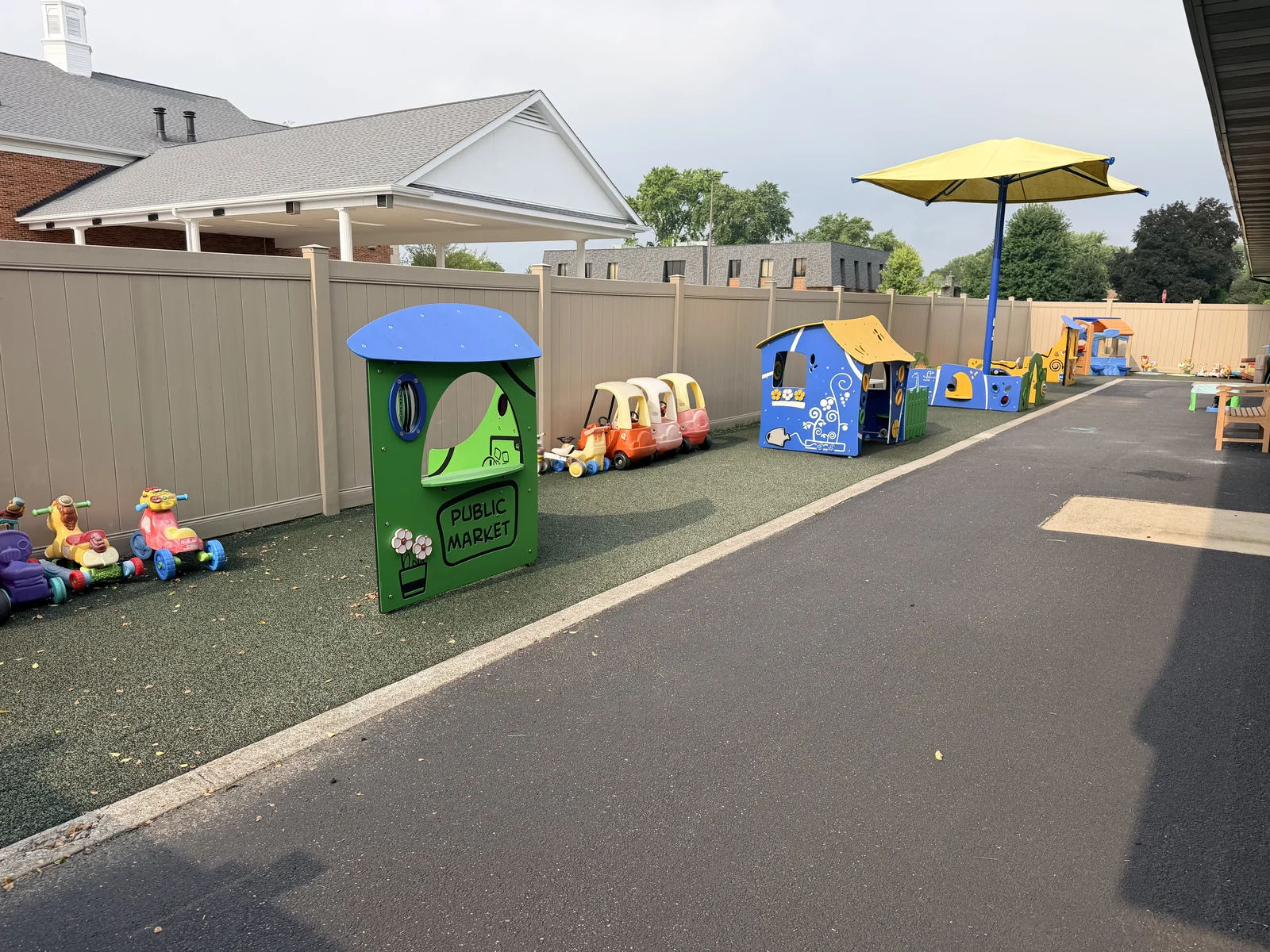 Separate toddler outdoor play area with playhouses, riding toys, a shaded table, and a fenced boundary